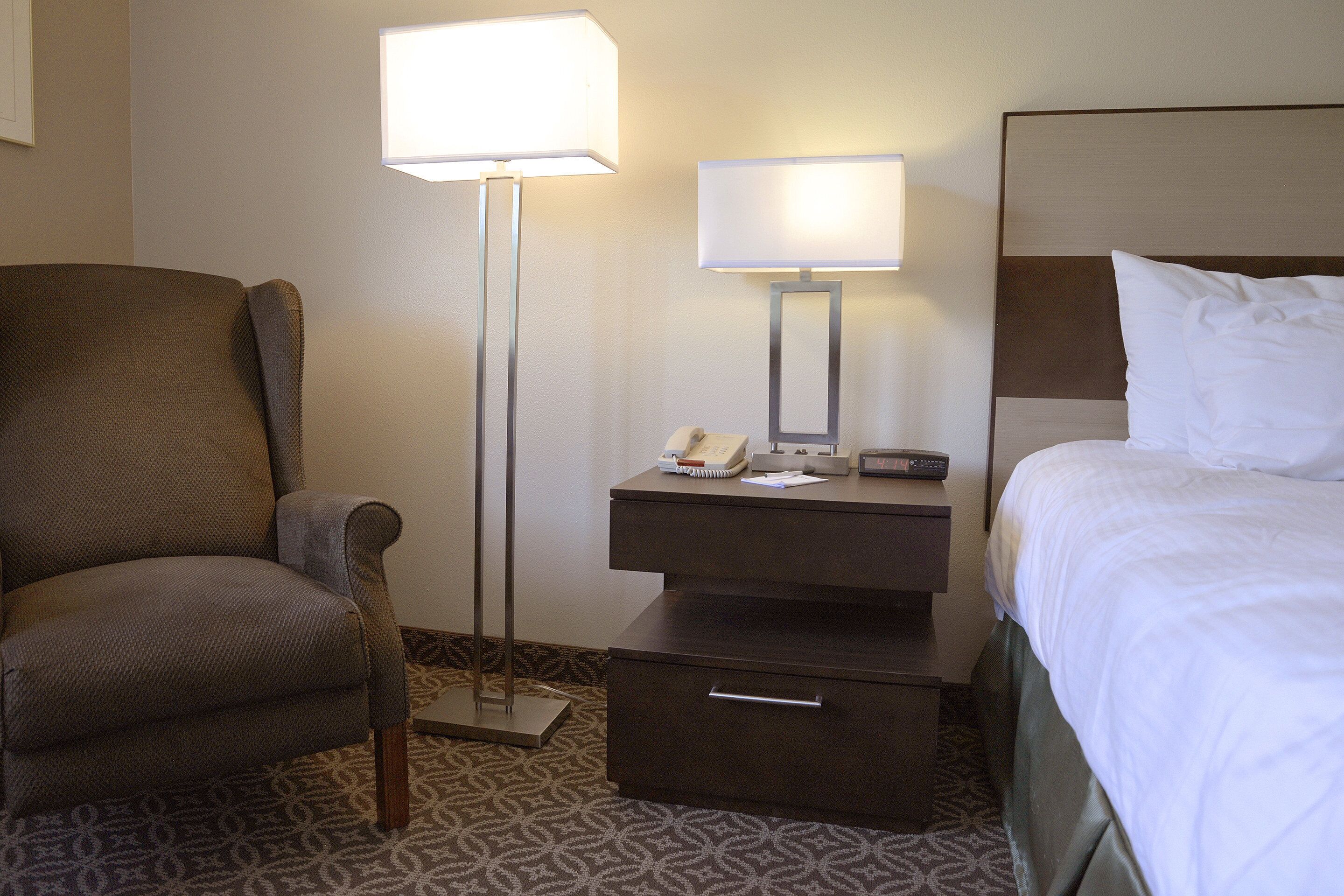 Una habitación de hotel con cama, una silla, una mesita de noche con lámpara y una mesita auxiliar con teléfono y reloj.