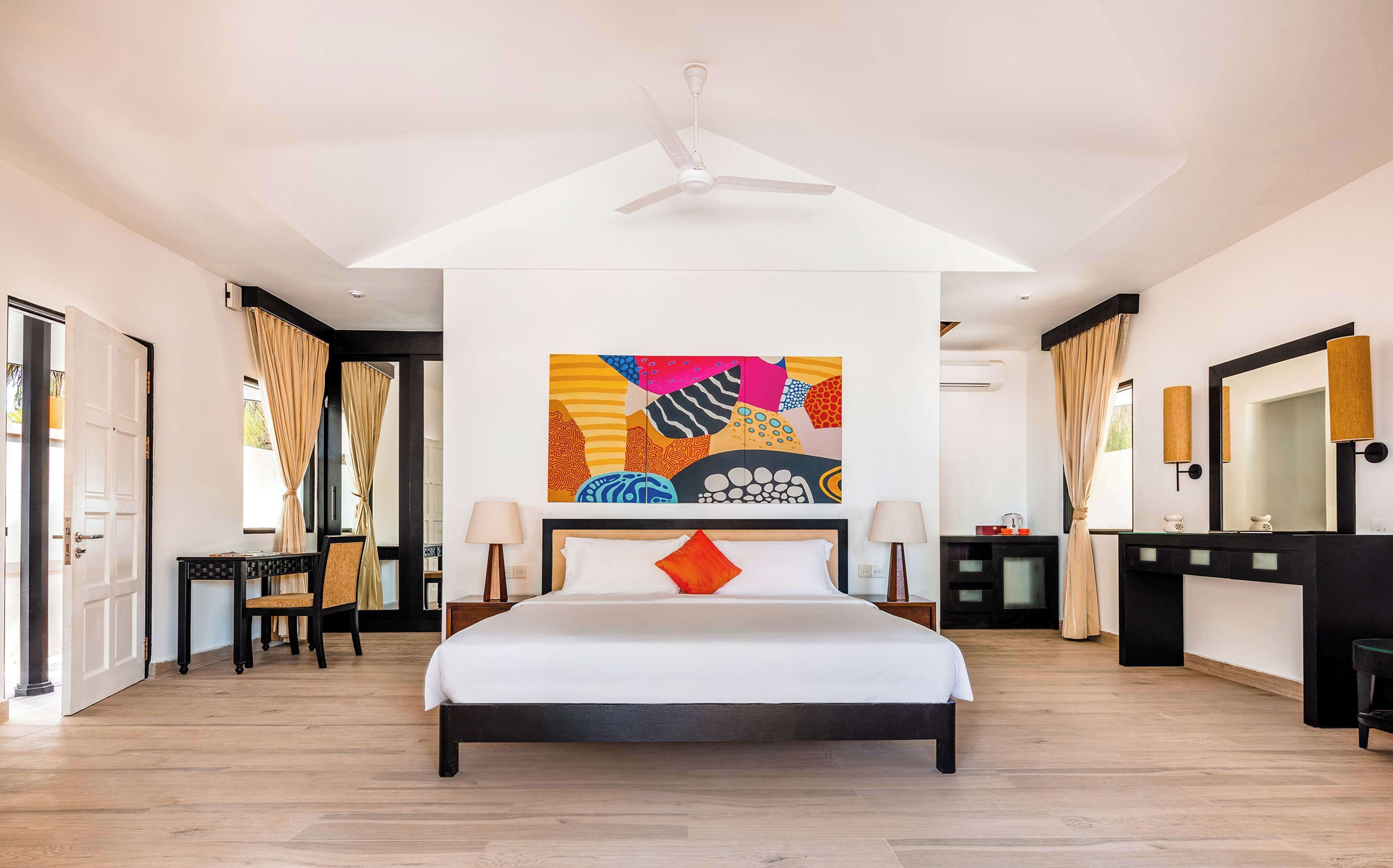 Um quarto espaçoso com uma cama grande, um ventilador de teto, um quadro abstrato colorido e um banheiro moderno com um espelho grande e um lavatório preto.