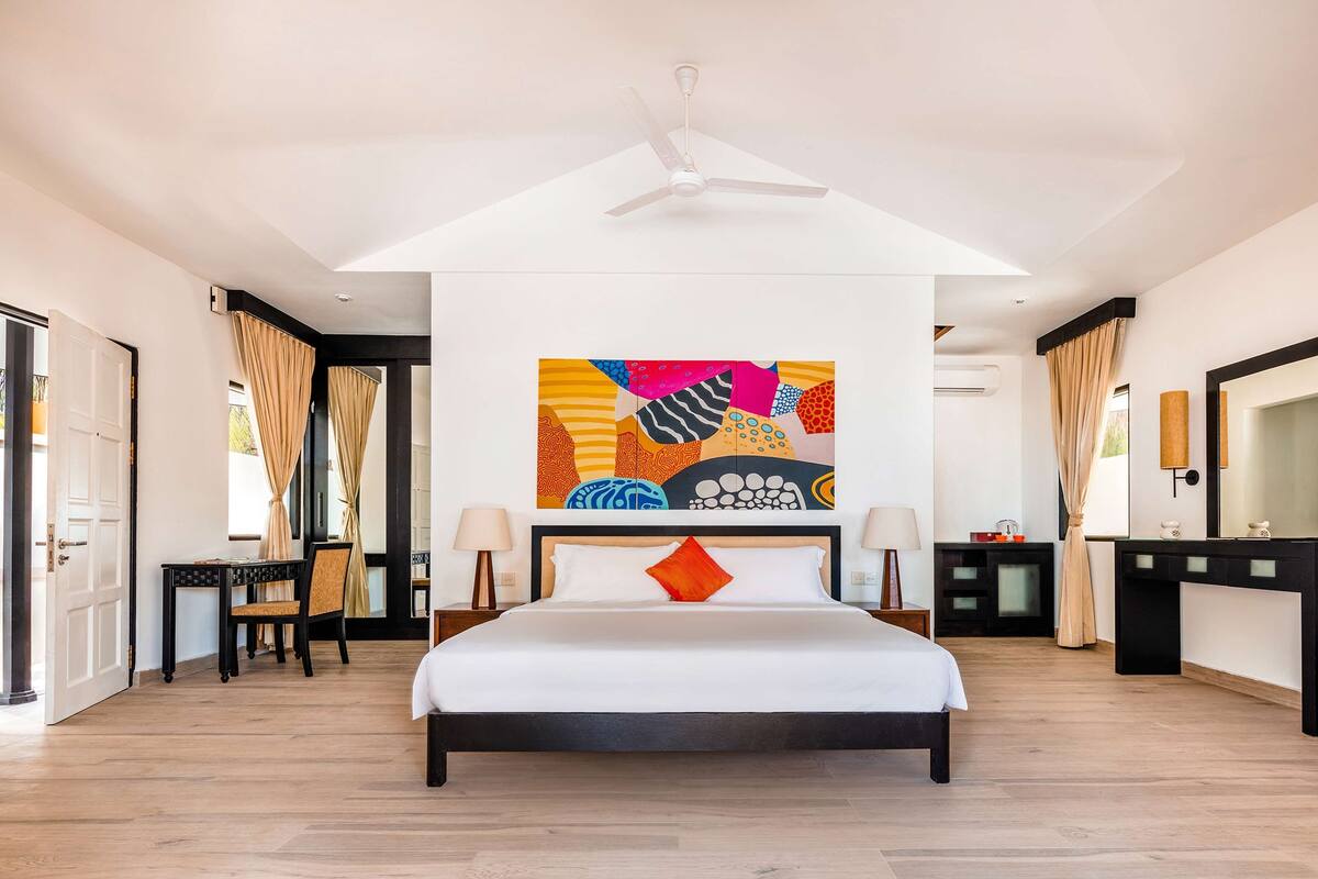 A spacious bedroom with a large bed, a ceiling fan, a colorful abstract painting, and a modern bathroom with a large mirror and a black vanity.