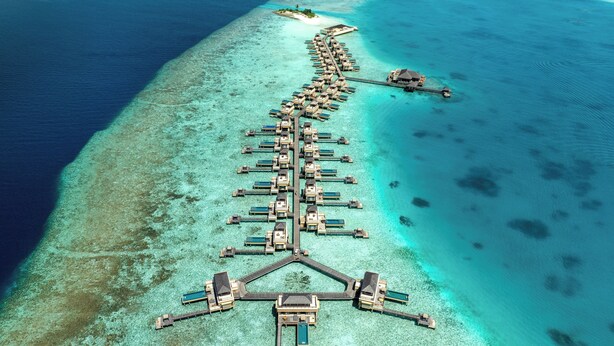 Eine Luftaufnahme eines Resorts mit überwasser Bungalows, die durch Gehwege verbunden sind, klarem türkisblauem Wasser und einer kleinen Insel mit einem Steg.