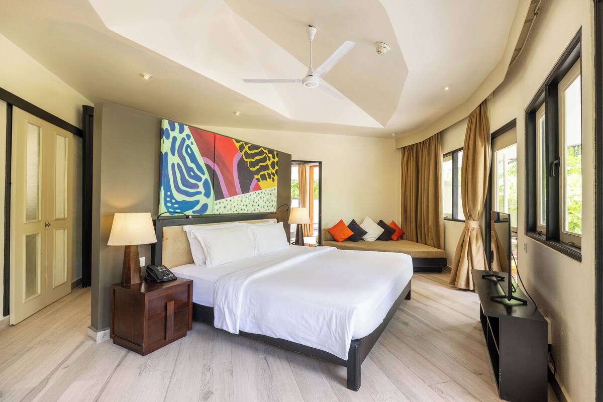 A modern hotel room with a large bed, a ceiling fan, a painting above the bed, and a TV stand.