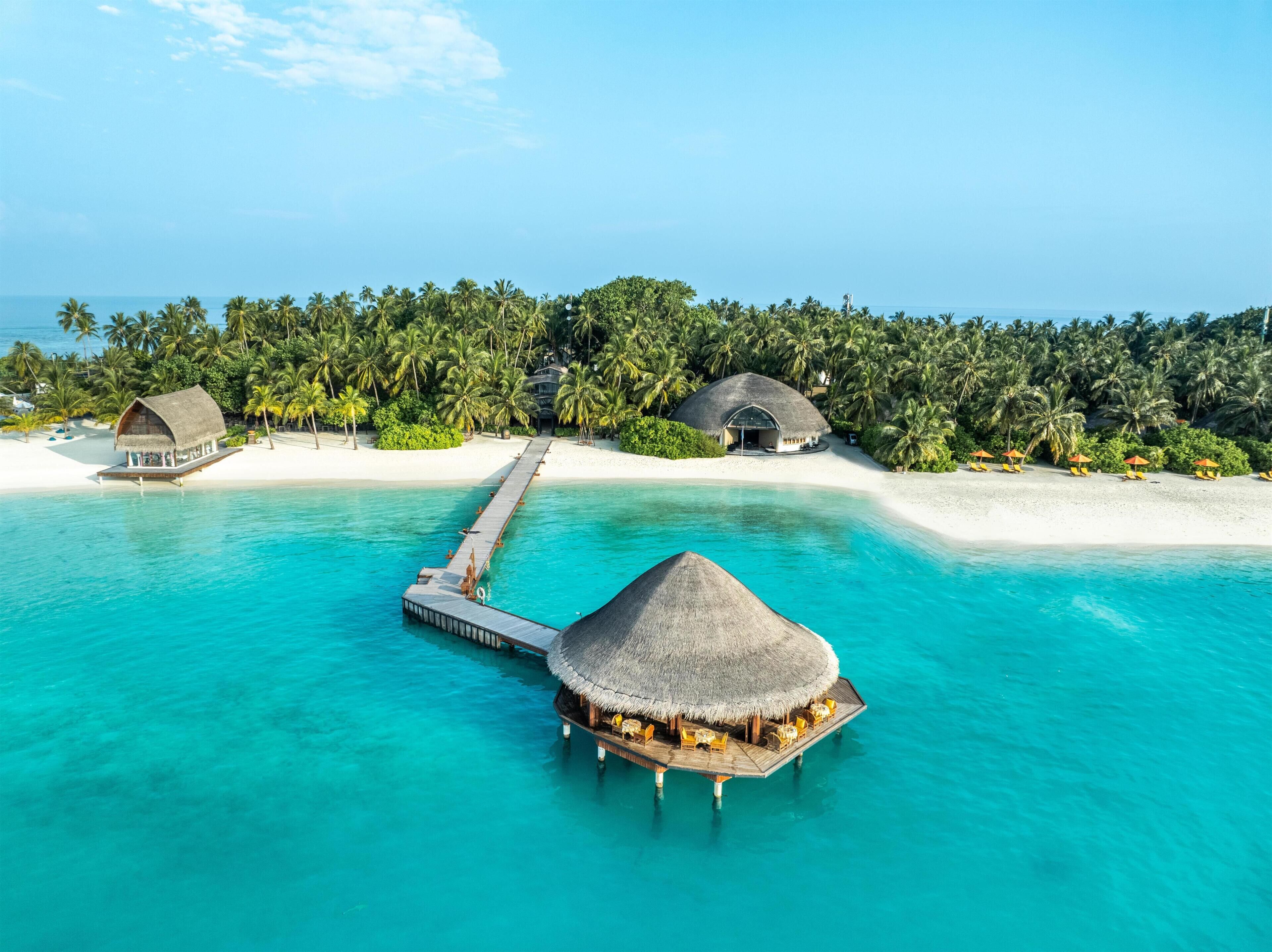 Ein Bungalow auf Stelzen mit einem Reetdach, eine Anlegestelle zu einem weißen Sandstrand und im Hintergrund üppige Palmen.
