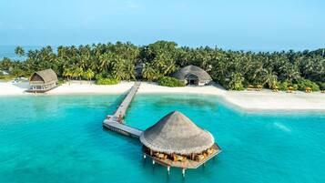 Ein Bungalow auf Stelzen mit einem Reetdach, eine Anlegestelle zu einem weißen Sandstrand und im Hintergrund üppige Palmen.