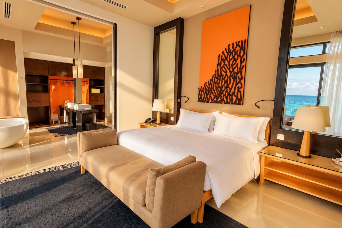A hotel room with a large bed, a sitting area, a bathtub, and a view of the sea.