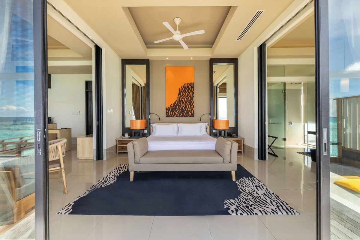 A modern bedroom with a large bed, a ceiling fan, and a view of the ocean through glass doors.
