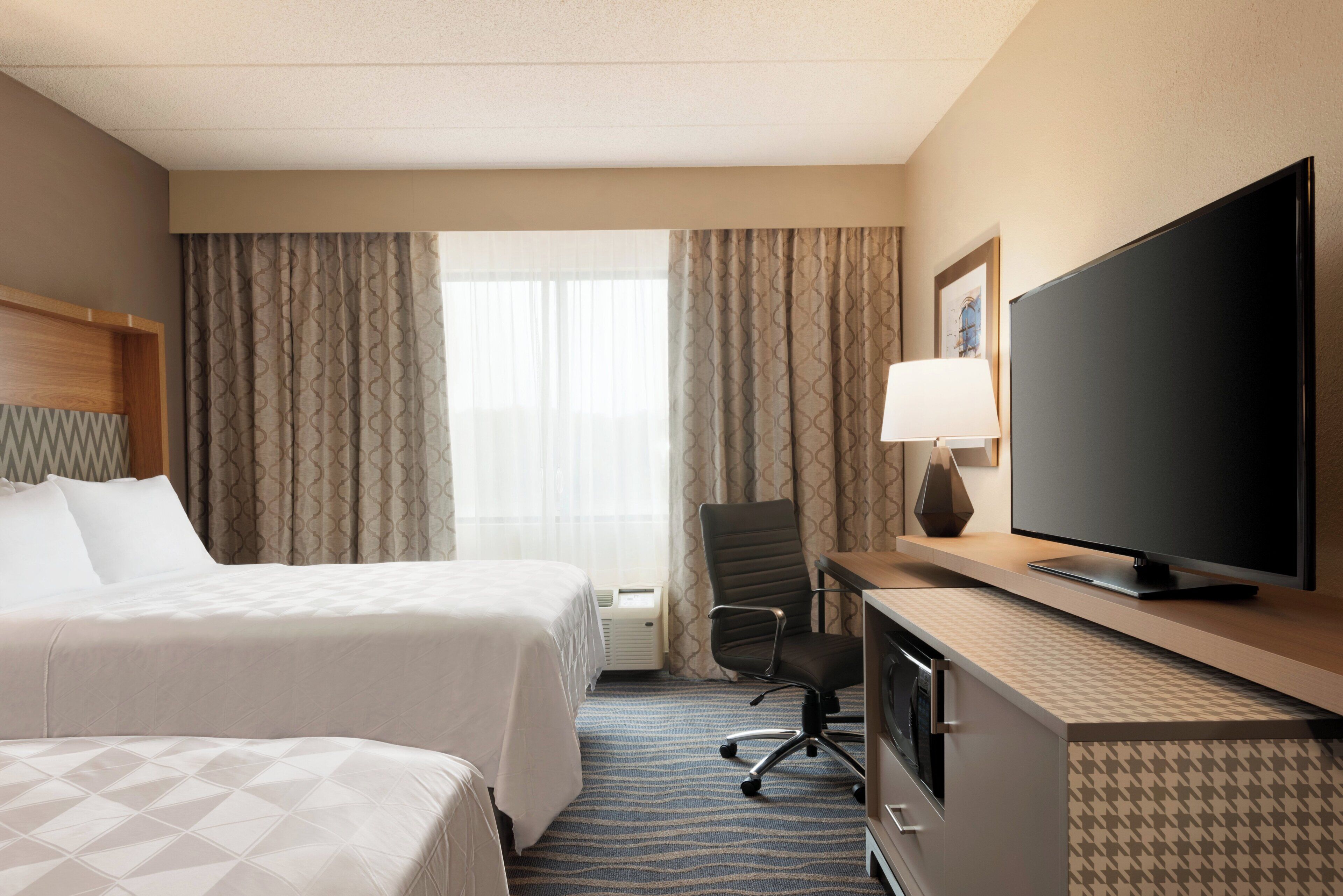 Une chambre d’hôtel avec un lit, un bureau avec une télévision, une chaise et une fenêtre avec des rideaux.