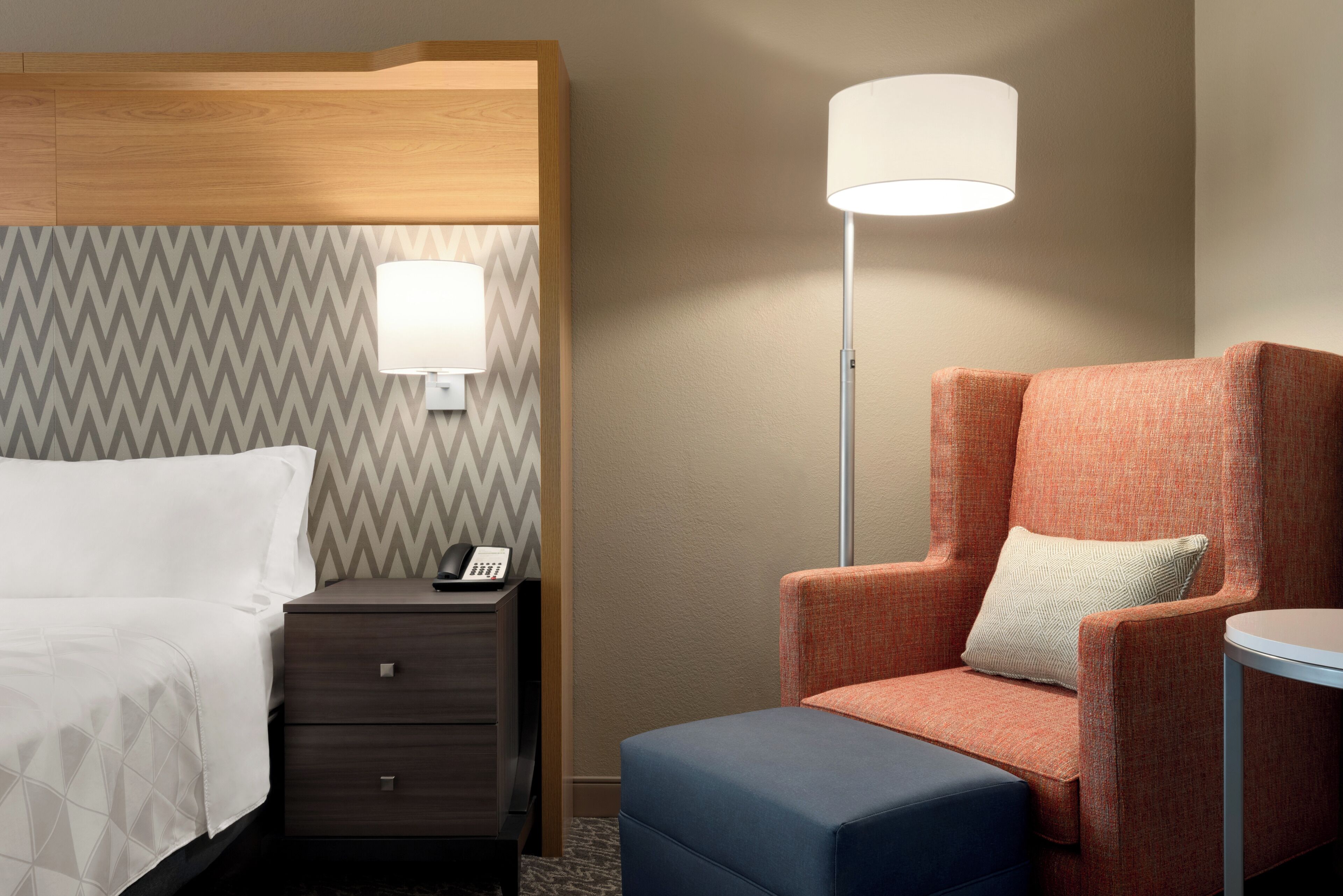 A hotel room with a bed, a nightstand with a phone, a lamp, an orange armchair, and a small table.