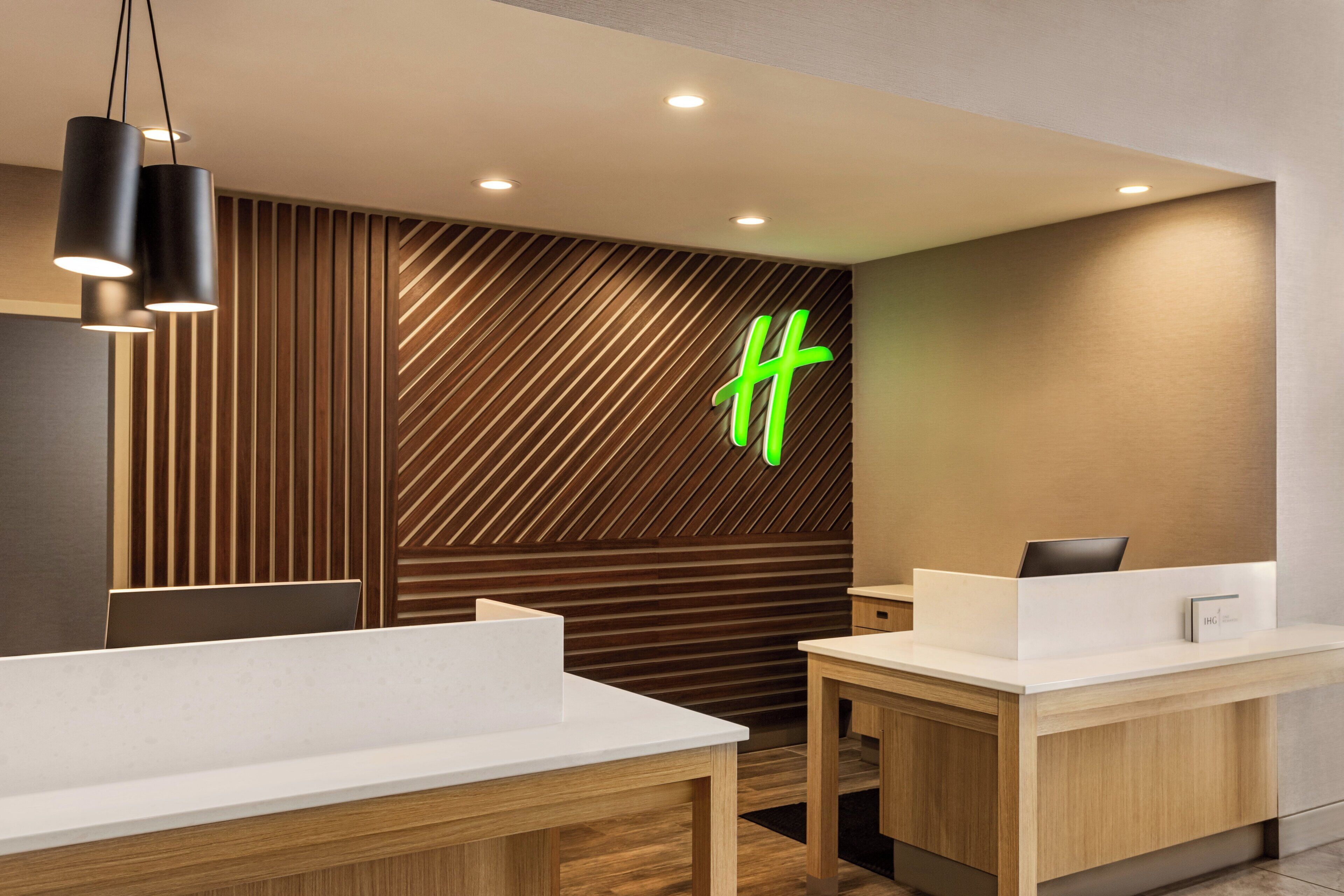 A modern hotel reception area with a wooden panel wall, a green 'H' sign, and a reception desk.
