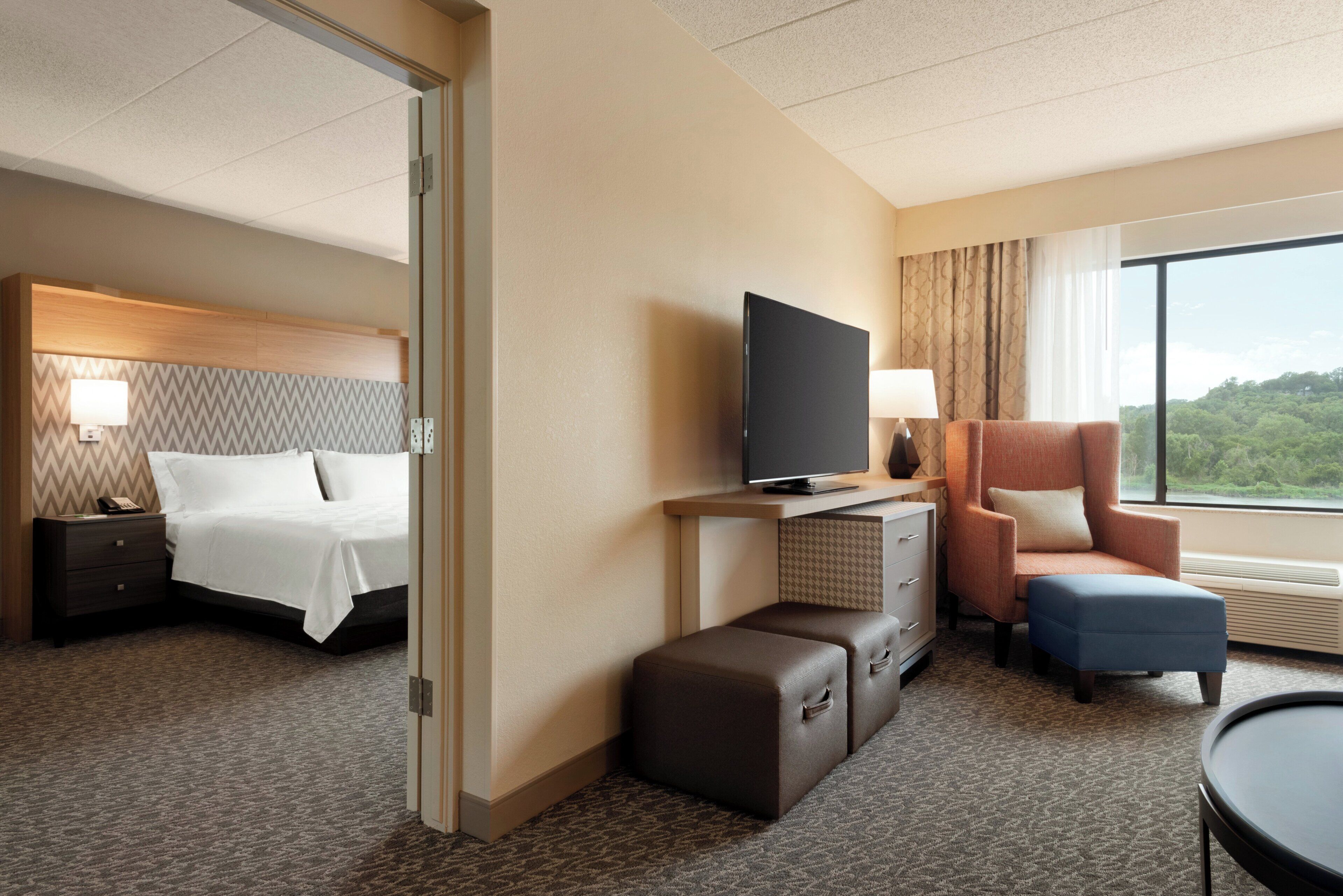 A hotel room with a bed, television, seating area, and a view of greenery.