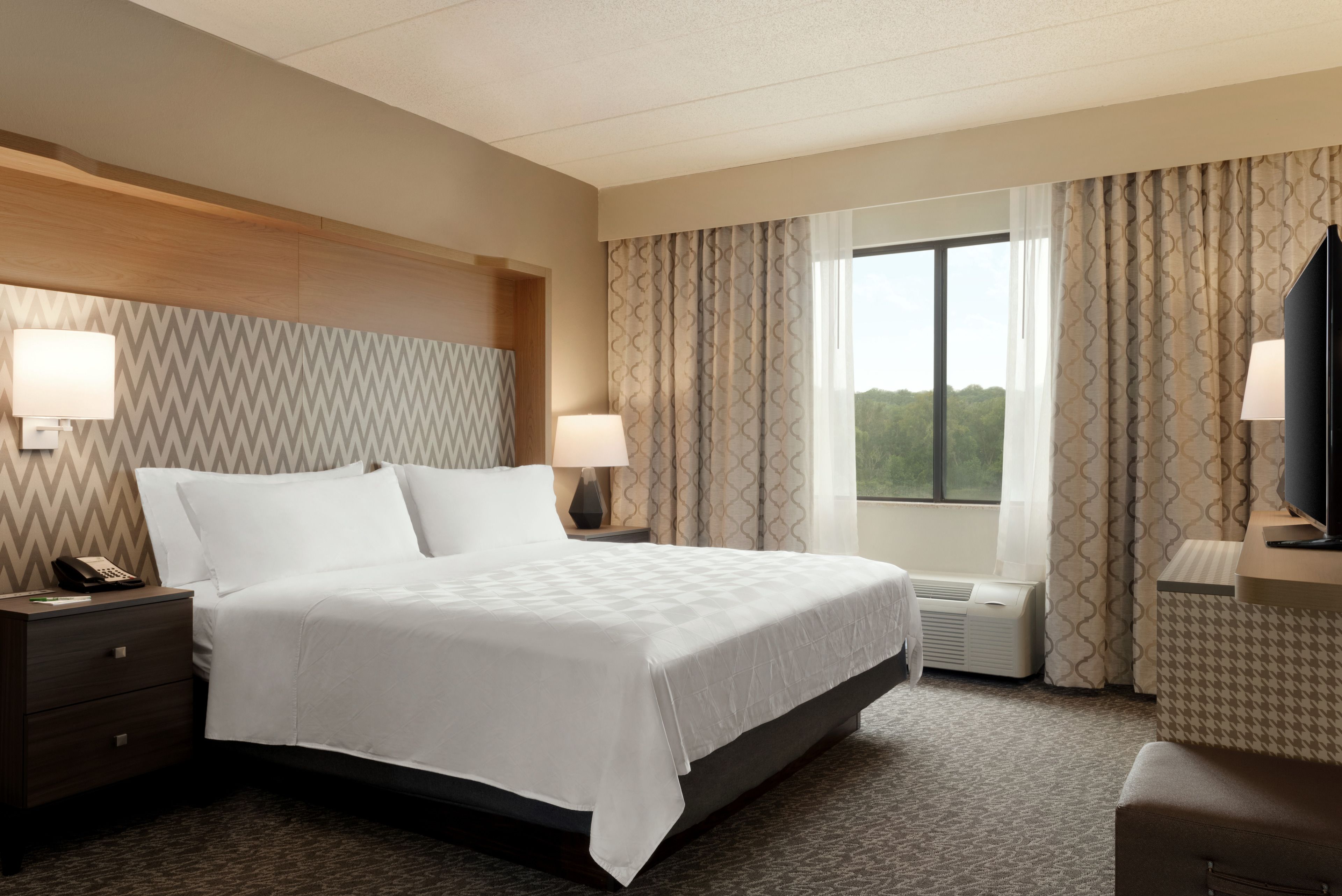 A hotel room with a bed, nightstands, lamps, a desk, and a window with curtains.