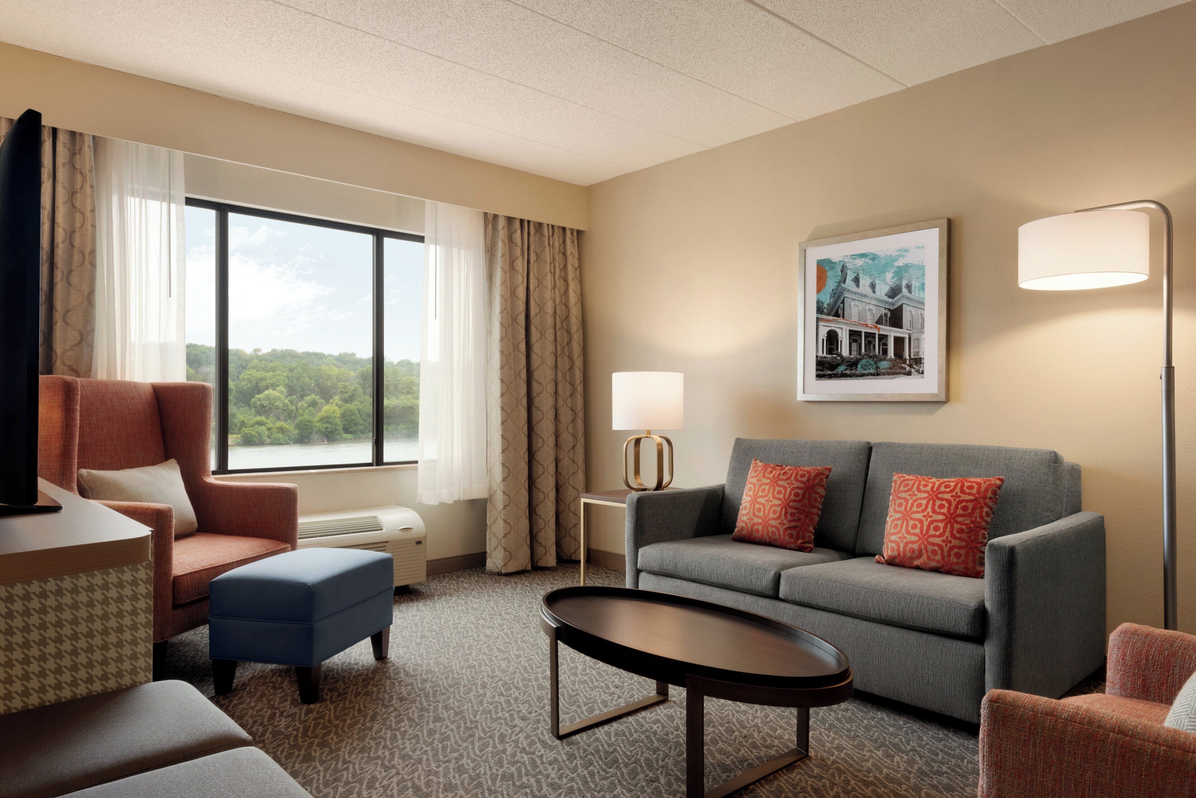 Habitación de hotel con sofá, sillón, reposapiés y una mesa. Se ve una ventana con cortinas, una lámpara de pared y un cuadro enmarcado.