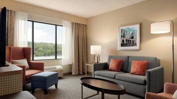 Habitación de hotel con sofá, sillón, reposapiés y una mesa. Se ve una ventana con cortinas, una lámpara de pared y un cuadro enmarcado.