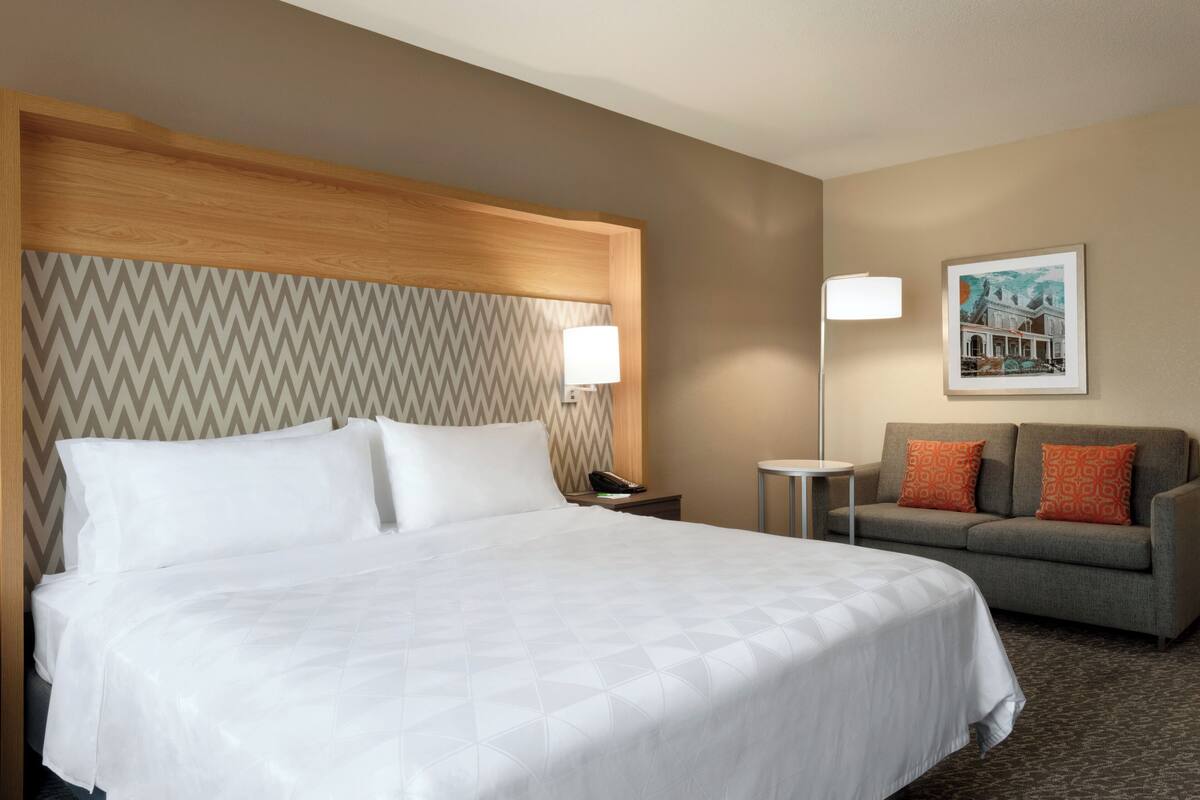 A hotel room with a large bed, a grey sofa, a small table, a nightstand, a lamp, and a framed picture on the wall.