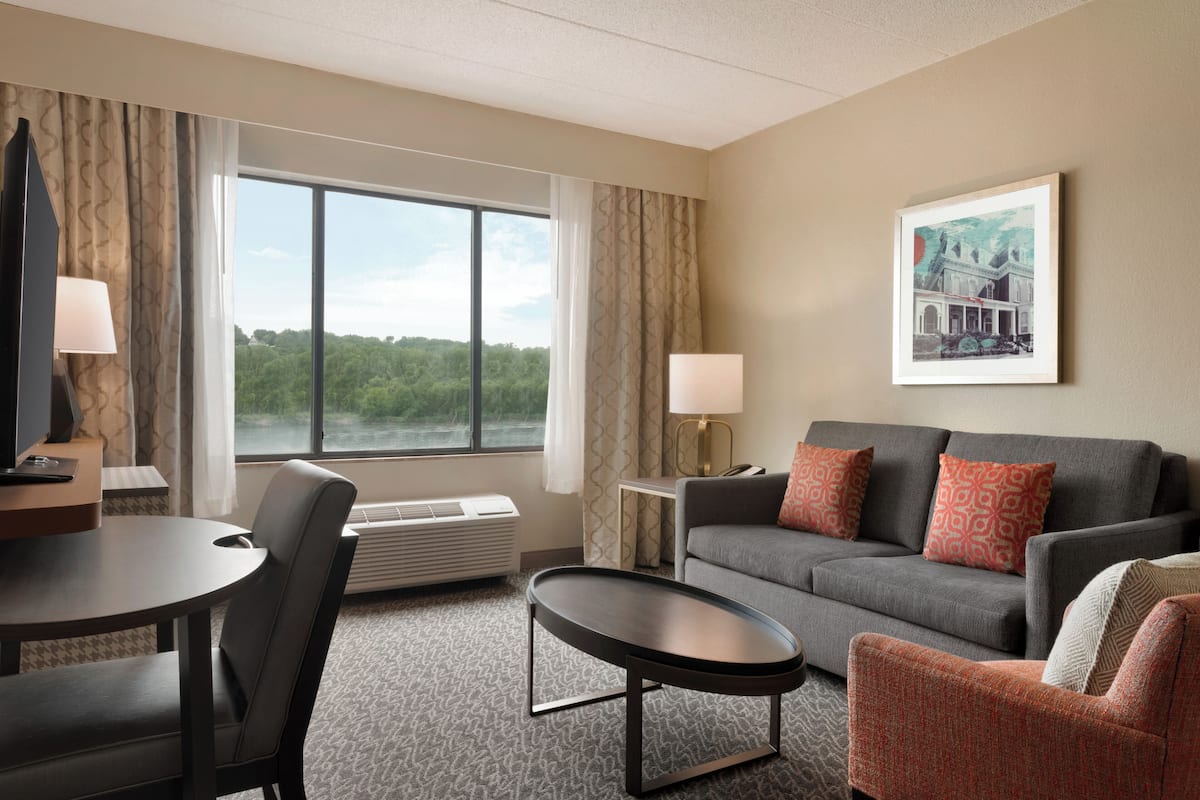 Une chambre d’hôtel avec un canapé gris, une petite table ronde, une chaise, un bureau avec une lampe et une grande fenêtre donnant sur un paysage verdoyant.