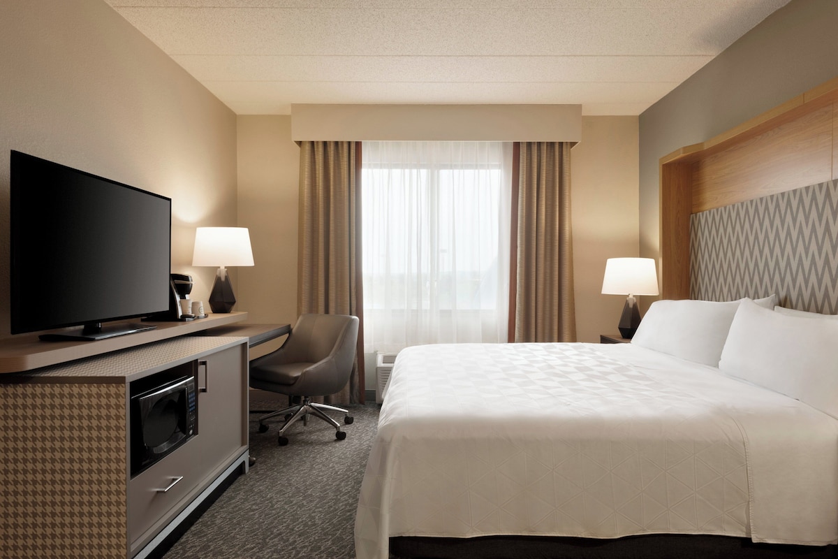 A hotel room with a bed, TV, desk, chair, lamps, and a window with curtains.