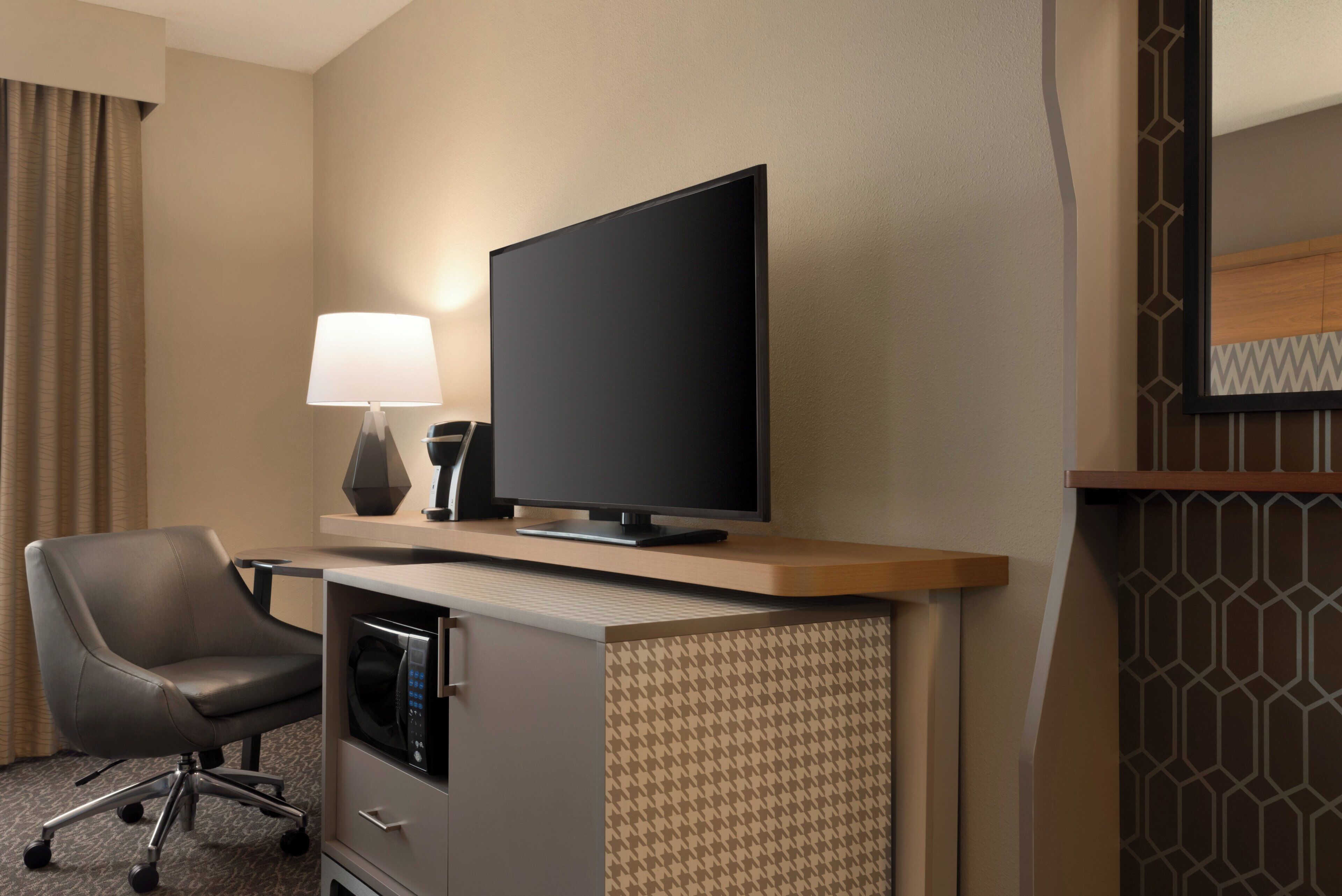 A hotel room with a flat-screen TV on a wooden stand, a grey office chair, a desk with a lamp, and a microwave.