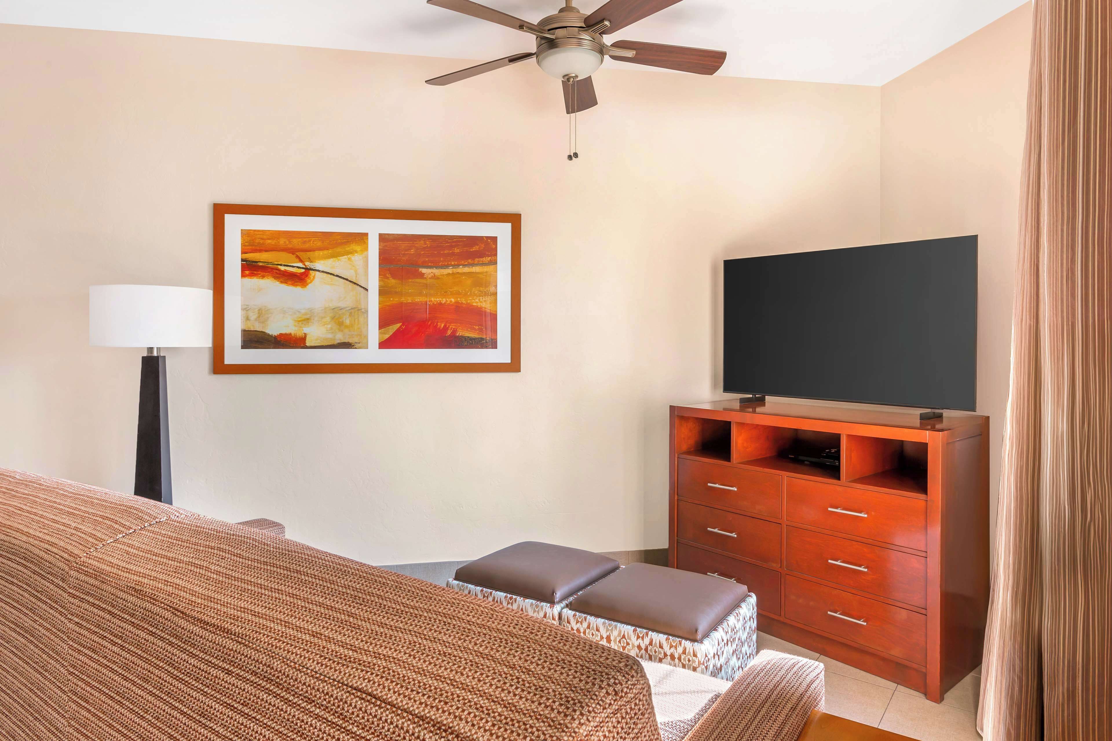 A hotel room with a bed, a TV on a wooden cabinet, a ceiling fan, and a painting on the wall.