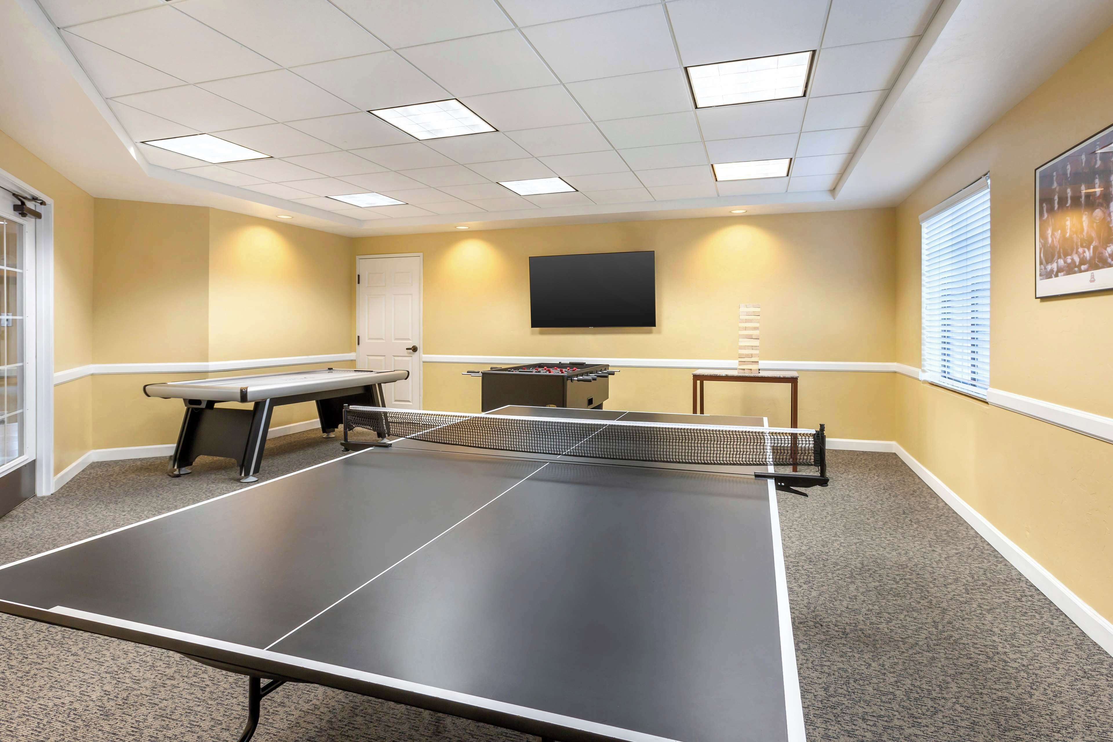 A room with a ping pong table, a bench, a TV, and a window with blinds.