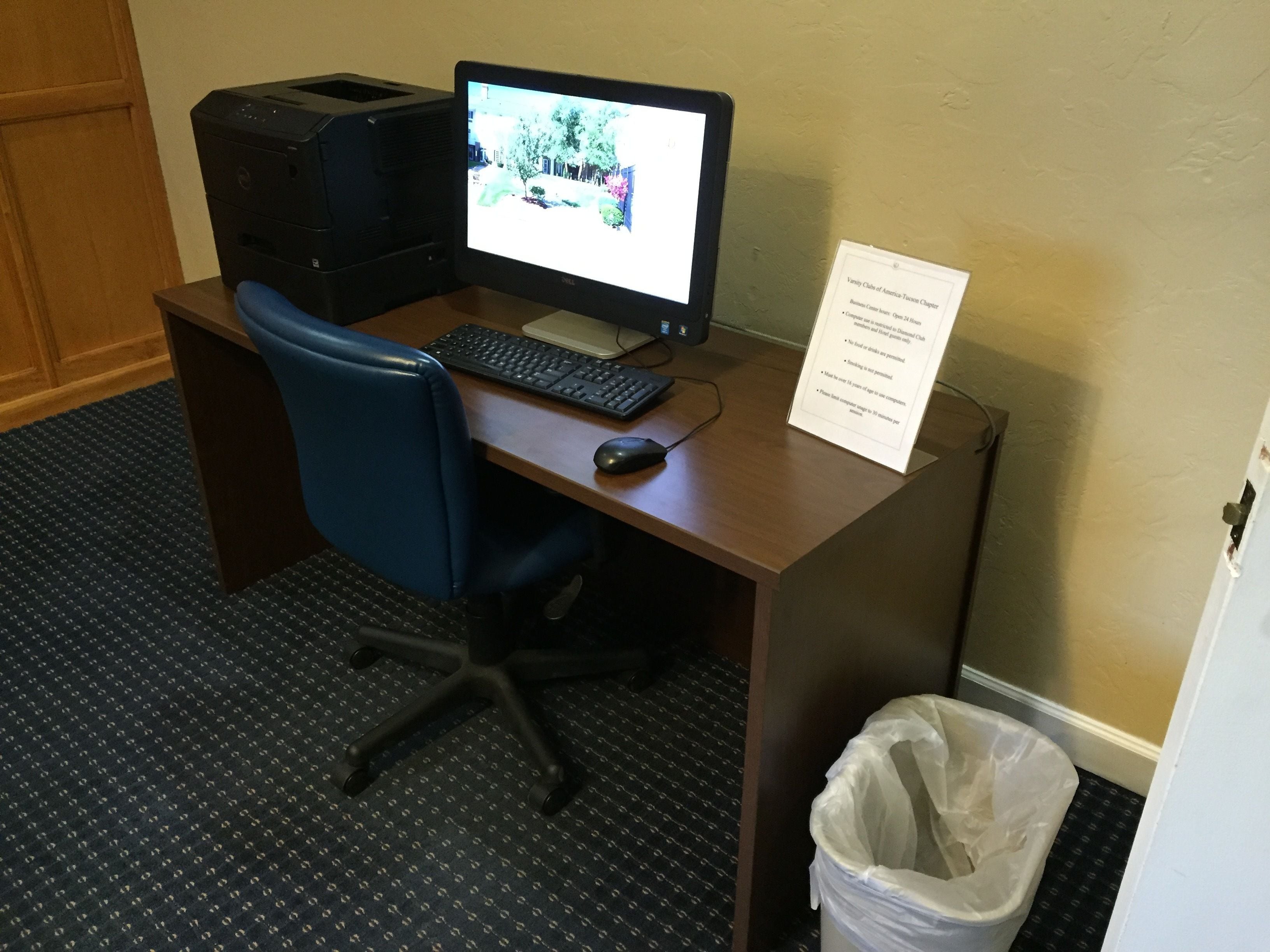 A hotel workspace with a desk, computer, keyboard, mouse, chair, and a trash bin.