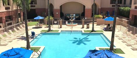 A hotel pool area with a clear blue pool, lounge chairs, umbrellas, and a building with a red-tiled roof.