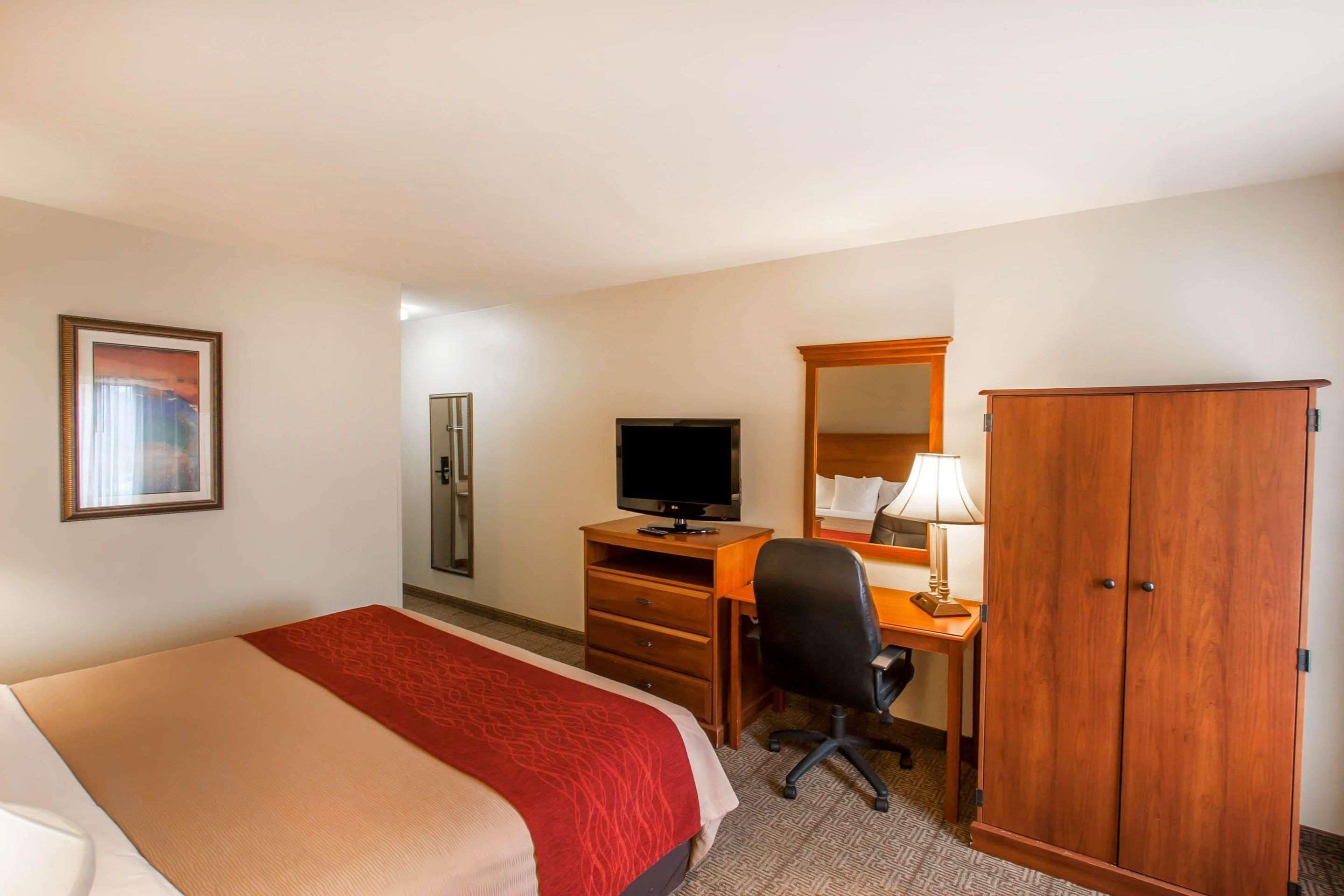 Una habitación de hotel con una cama, un escritorio con computadora, televisor, armario y un cuadro en la pared.