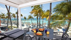 A table with breakfast items, a glass of orange juice, and a view of the beach and palm trees. - Divi Aruba Phoenix Beach Resort (Noord)