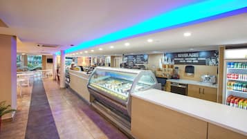Una cafetería con un expositor de vidrio, una carta de bebidas y una zona de estar con mesas y sillas.