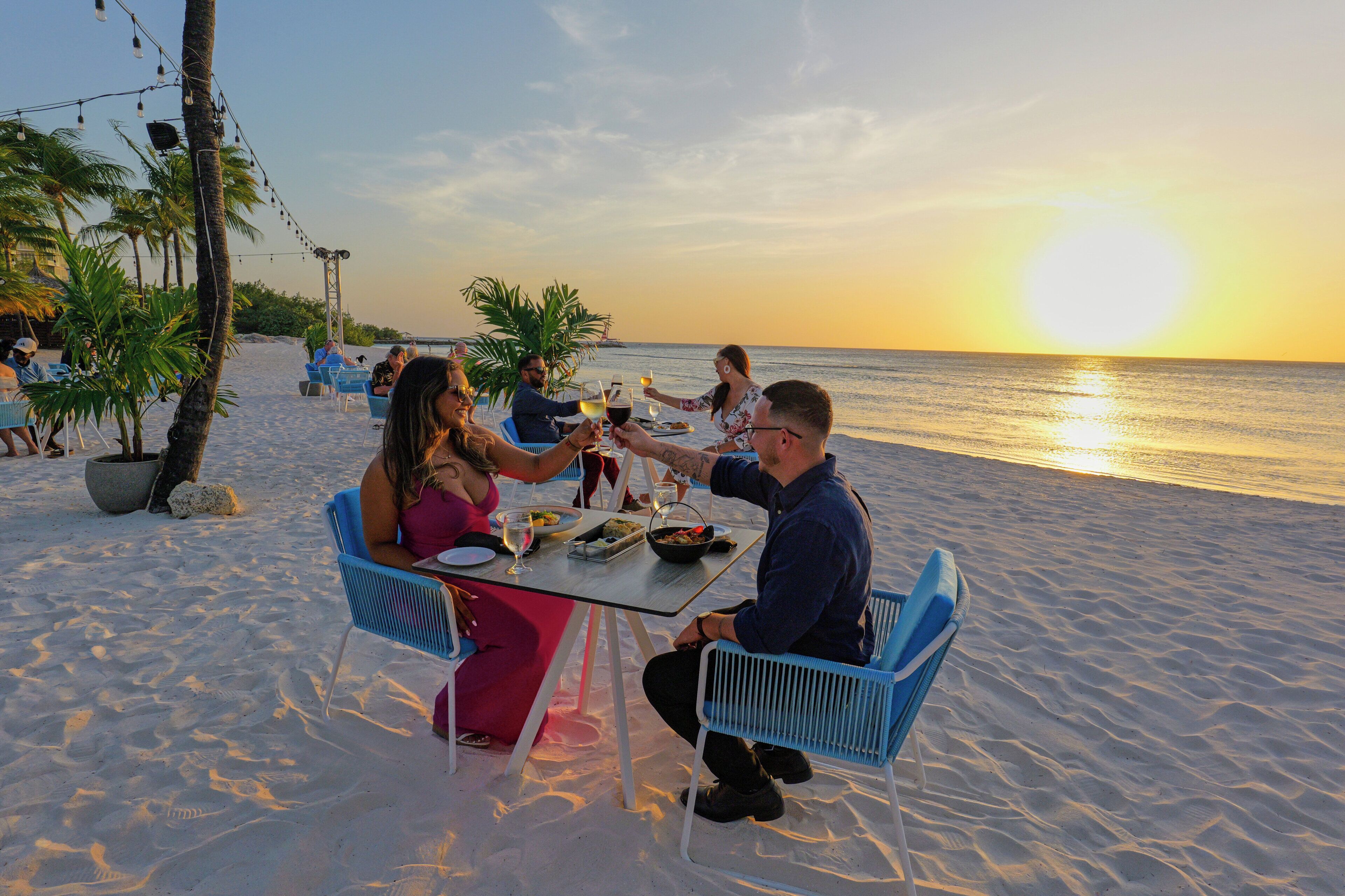 Un área de comedor frente a la playa, con mesas y sillas, palmeras y vistas al océano.