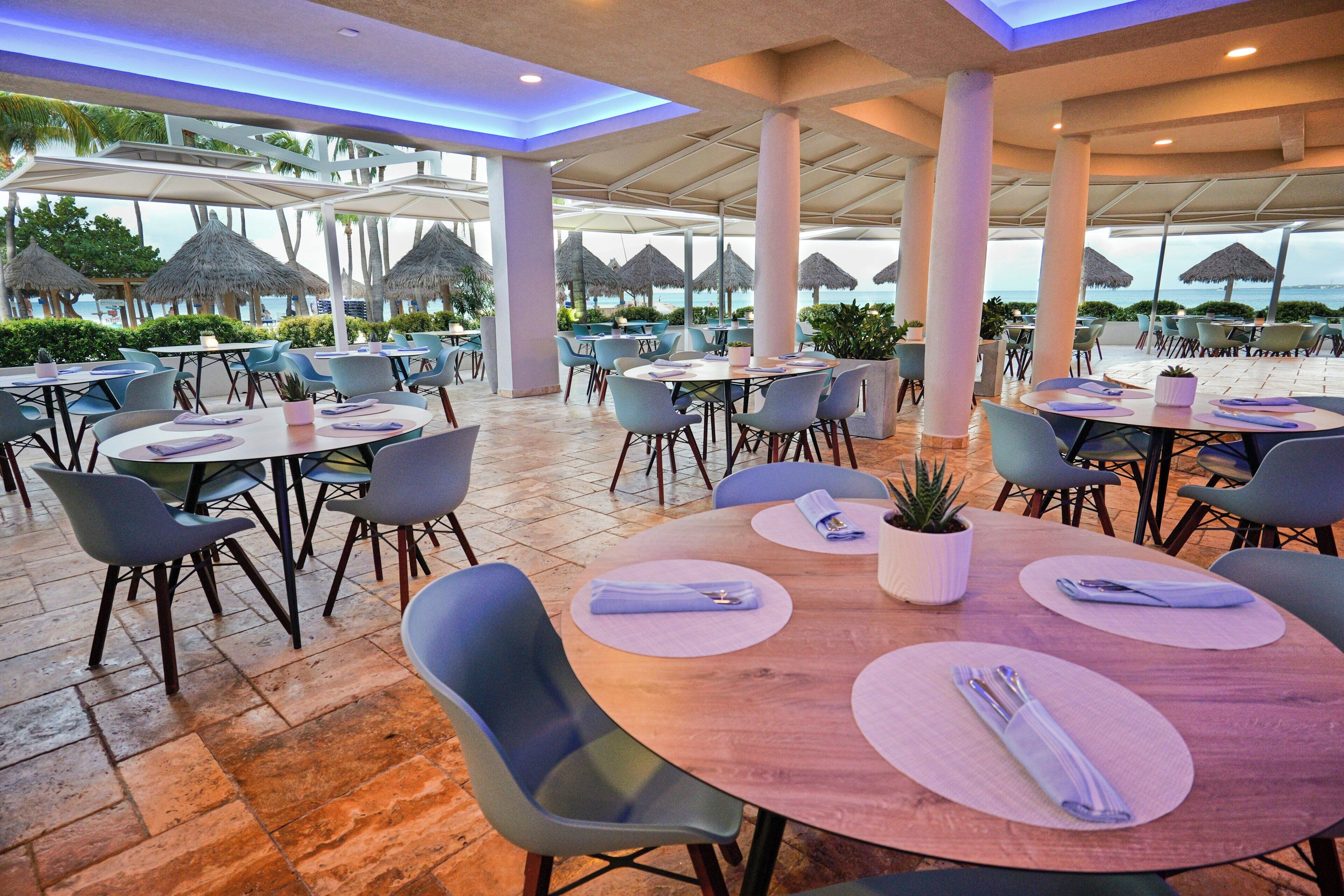An outdoor dining area with round tables, chairs, and umbrellas, overlooking a beach.