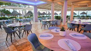 An outdoor dining area with round tables, chairs, and umbrellas, overlooking a beach.