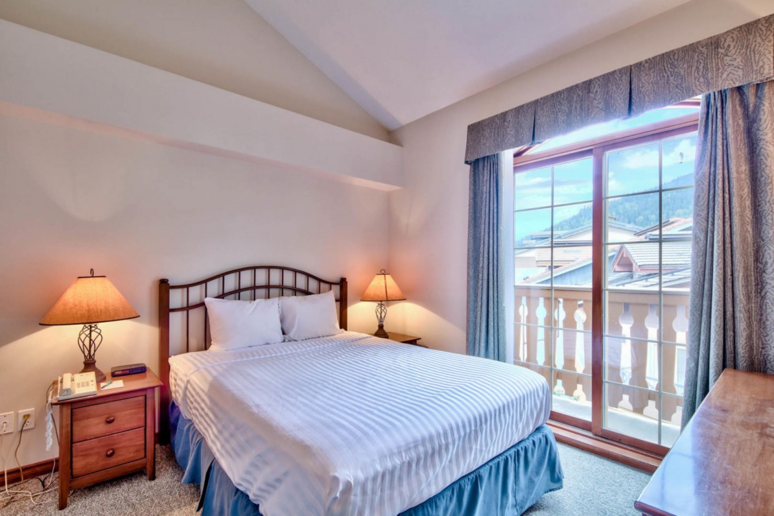 A hotel room with a large bed, two bedside tables with lamps, a desk, and a balcony with a view of buildings.