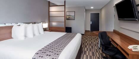 A hotel room with a large bed, a desk with a chair, a television, and a door leading to another room.
