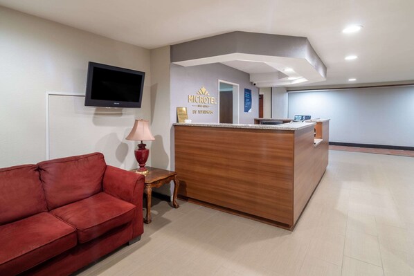 Un espace d’accueil d’hôtel avec un comptoir de réception en bois, un canapé rouge, un téléviseur fixé au mur et une lampe sur une petite table.