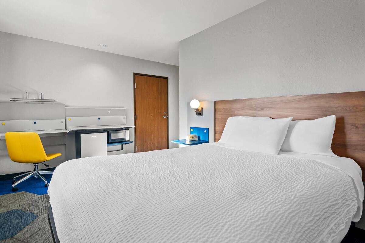 A hotel room with a large bed, a desk, a chair, and a nightstand with a lamp.