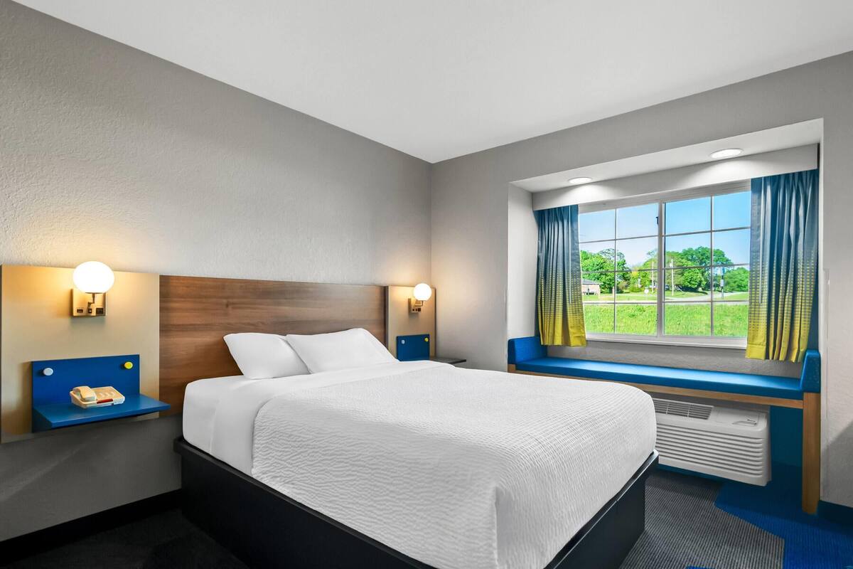 A hotel room with a bed, two bedside tables, a window with curtains, and a view of greenery outside.