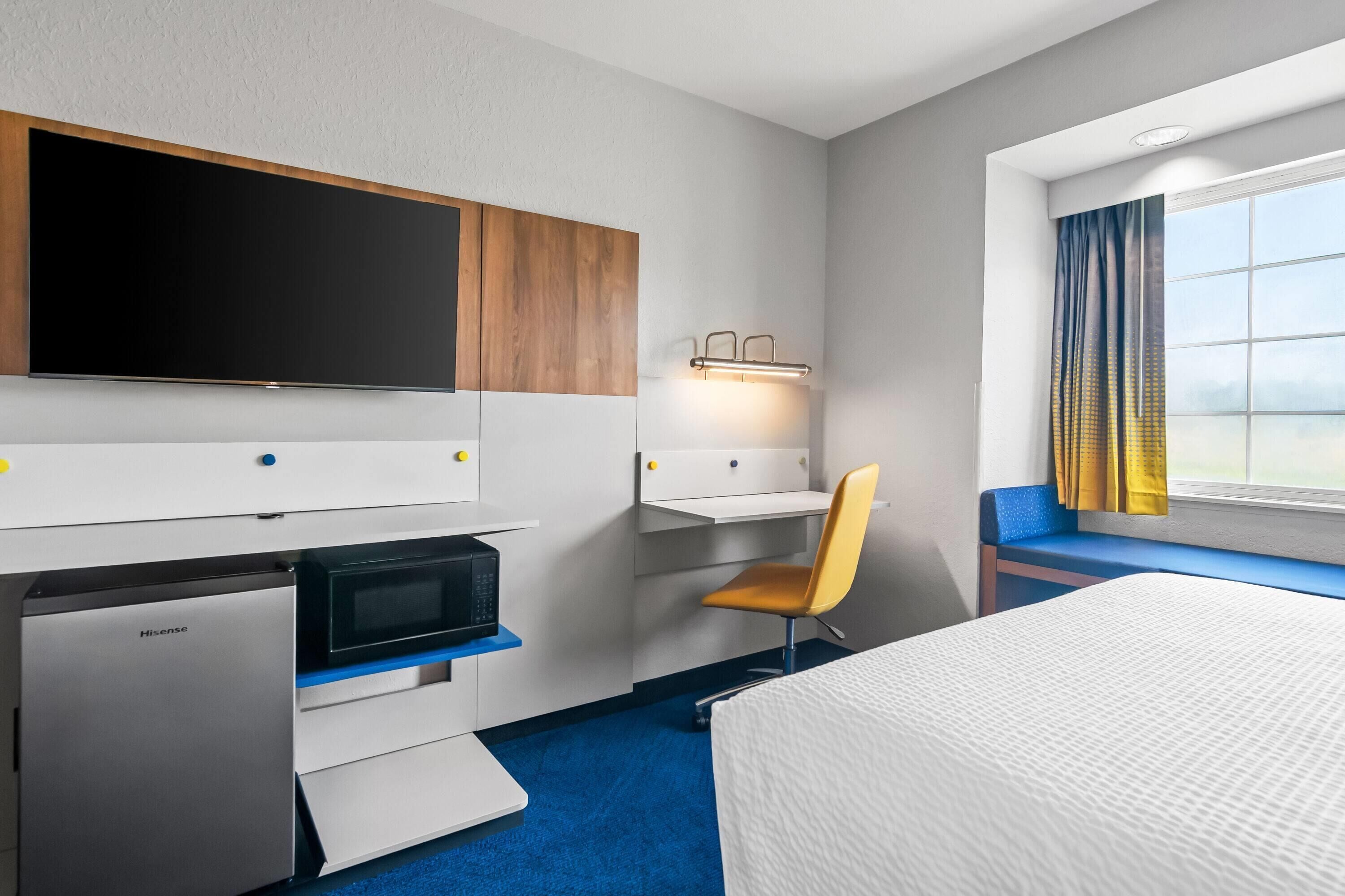 Une chambre d’hôtel équipée d’un lit, d’un bureau avec une chaise, d’une télévision, d’un four à micro-ondes et d’un petit réfrigérateur.