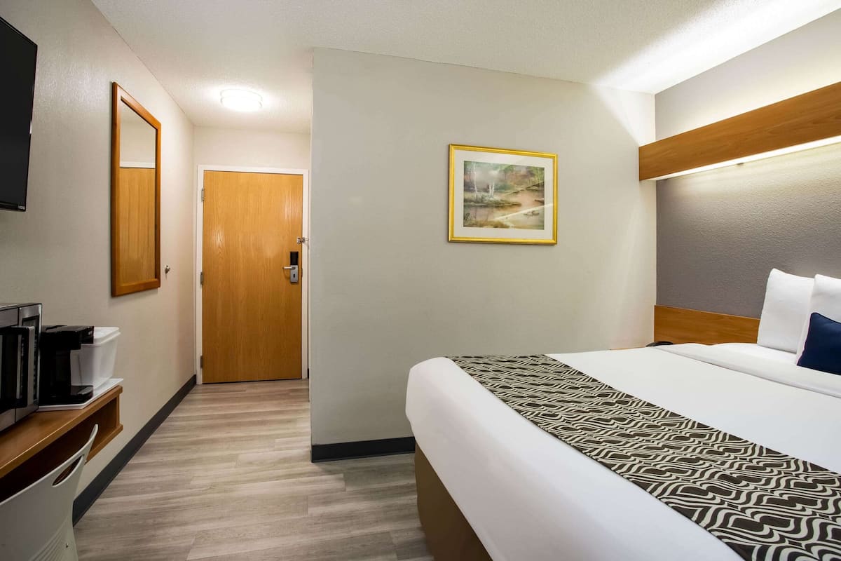 Habitación de hotel con cama, mesita de noche con cafetera, espejo, puerta y un cuadro en la pared.