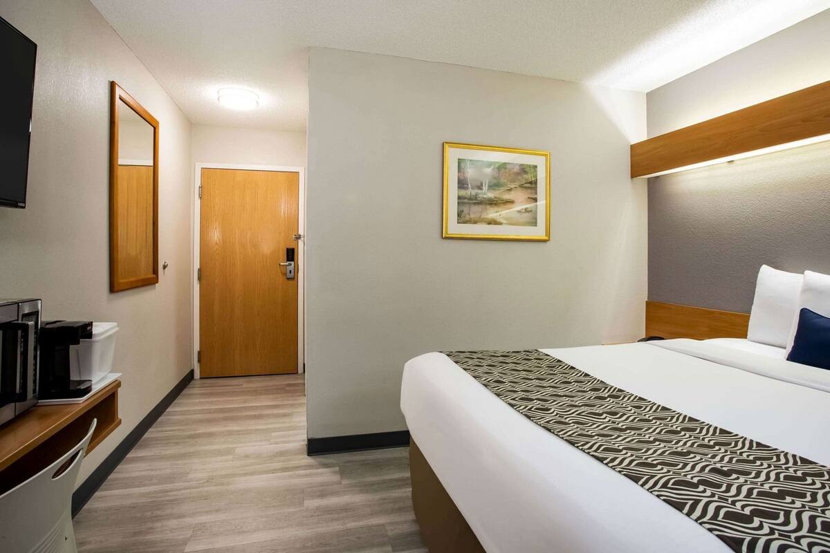 Una habitación de hotel con cama, televisión, cafetera, puerta y un cuadro en la pared.