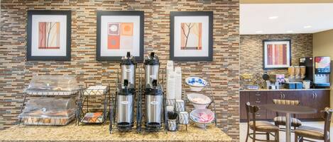 A hotel reception area with a brick-patterned wall, framed artwork, a coffee station with dispensers, and a seating area with a table and chairs.