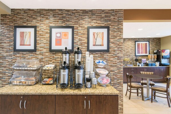 A hotel reception area with a brick-patterned wall, framed artwork, a coffee station with dispensers, and a seating area with a table and chairs.