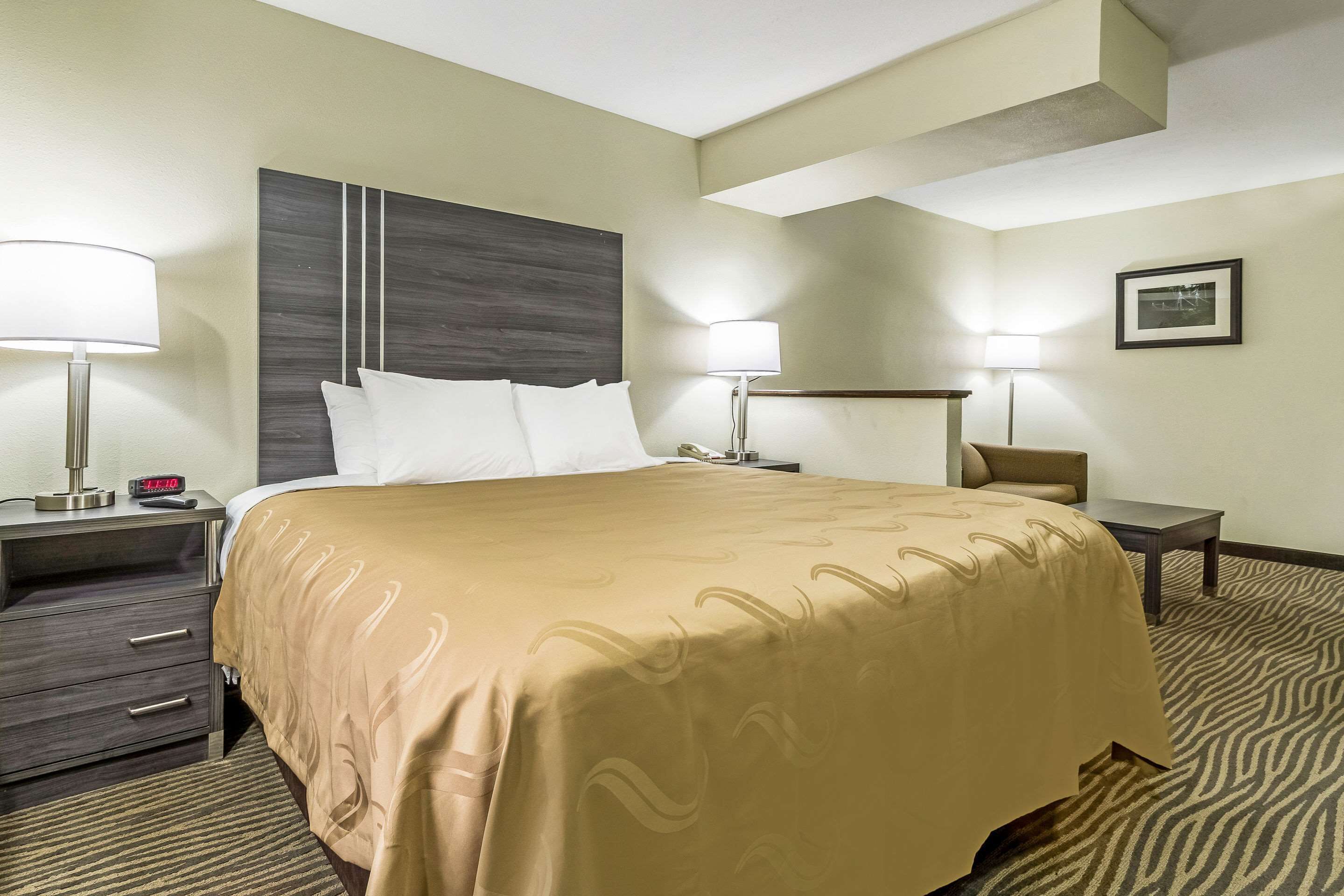 Habitación de hotel con una cama grande, dos mesitas de noche con lámparas, una mesita auxiliar, un reloj y un cuadro en la pared.