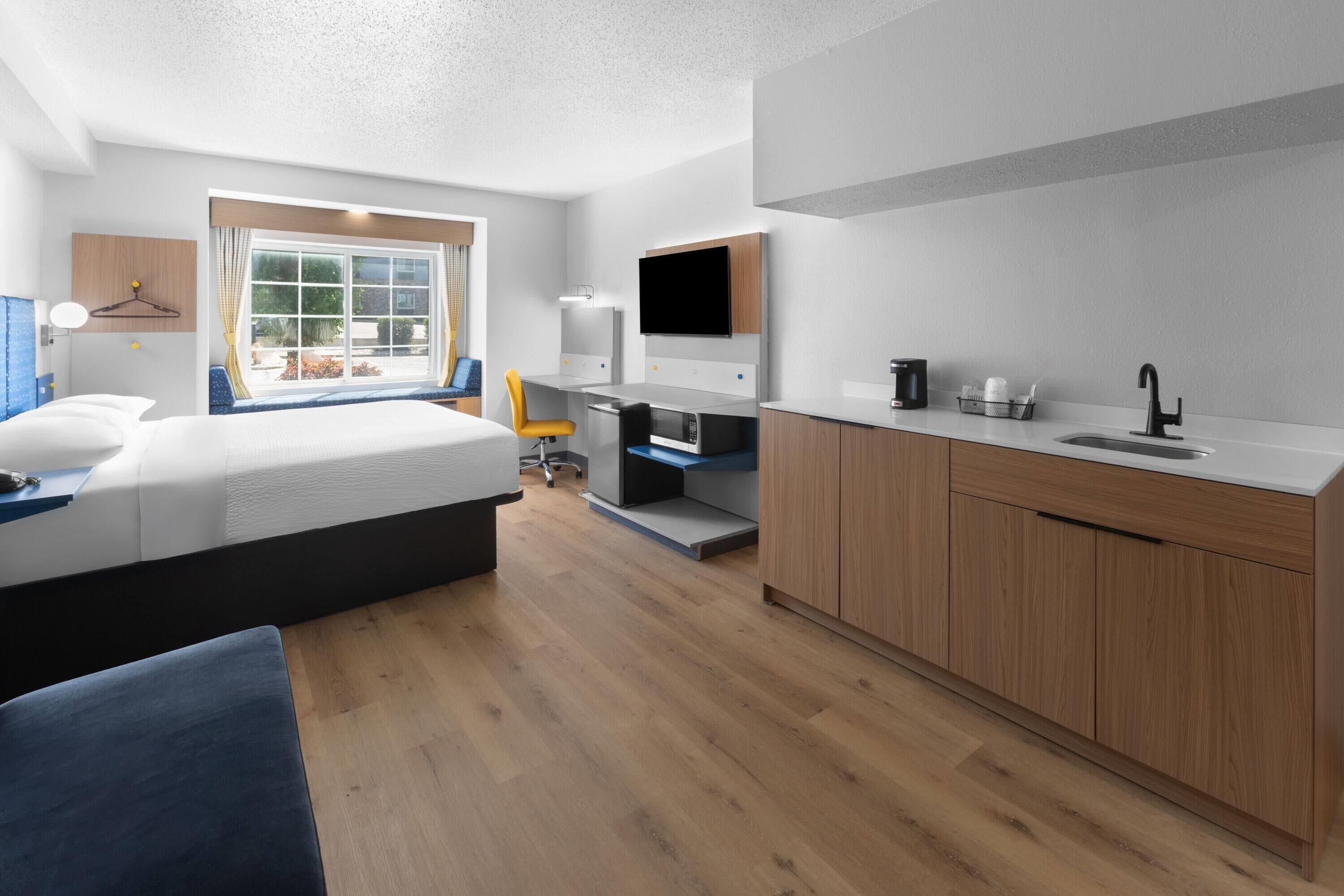 Quarto de hotel moderno com cama, escrivaninha, televisão e cozinha compacta.