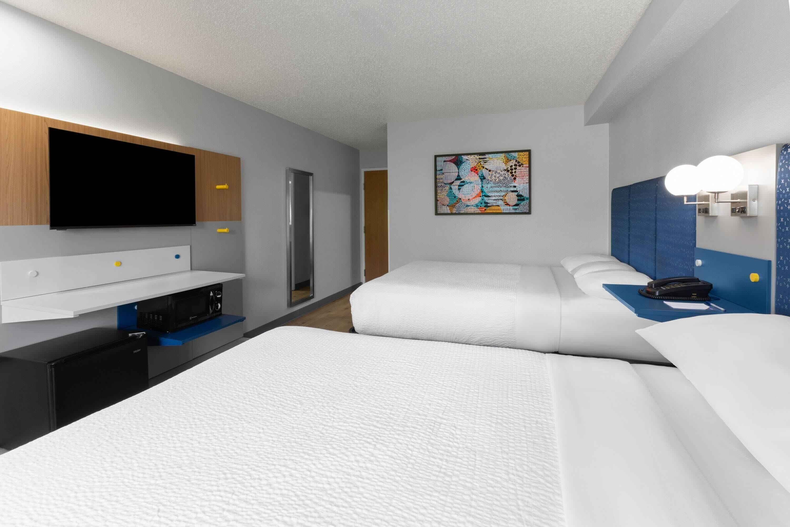 A modern hotel room with a flat-screen TV, a microwave, a mini-fridge, a desk with a computer, two beds, a painting, and a bathroom with a mirror and lights.