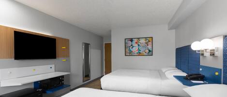 A modern hotel room with a flat-screen TV, a microwave, a mini-fridge, a desk with a computer, two beds, a painting, and a bathroom with a mirror and lights.