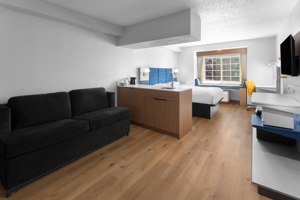 A modern hotel room with a black sofa, wooden flooring, a kitchenette, a bed with a blue headboard, and a window with a view of buildings.