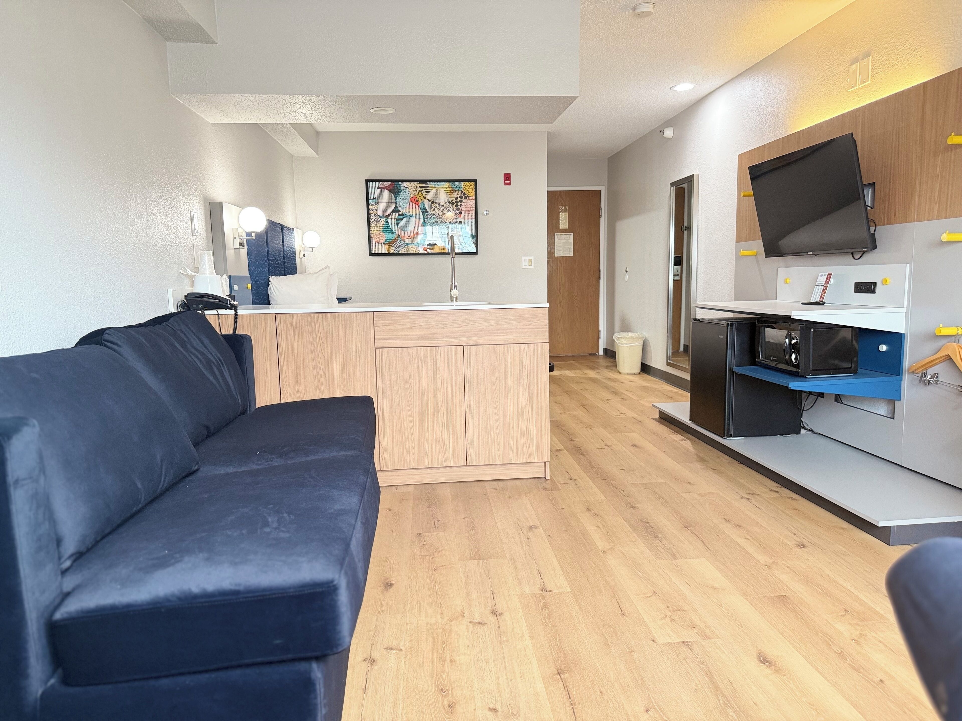 A modern living room with a dark blue sofa, a wooden cabinet, and a kitchen area with a microwave and a TV mounted on the wall.