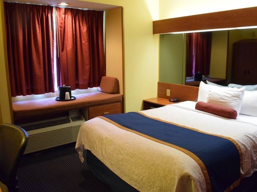 A hotel room with a bed, a desk with a chair, a window with curtains, a nightstand, and a mirror.