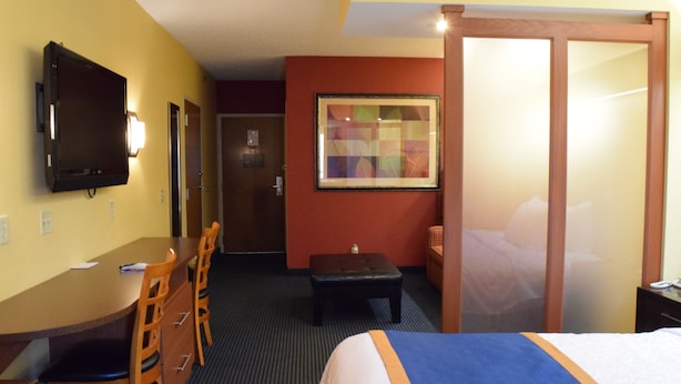 A hotel room with a bed, a desk with chairs, a television, a framed picture on the wall, and a door.