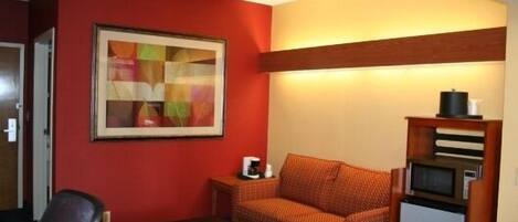 A compact hotel room with a red accent wall, a framed abstract painting, a small sofa, a coffee table, and a compact kitchenette with a microwave.