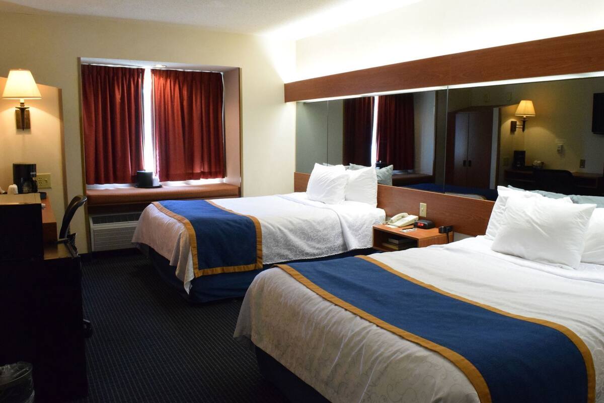 A hotel room with two beds, a desk, a chair, a lamp, a mirror, and a window with curtains.